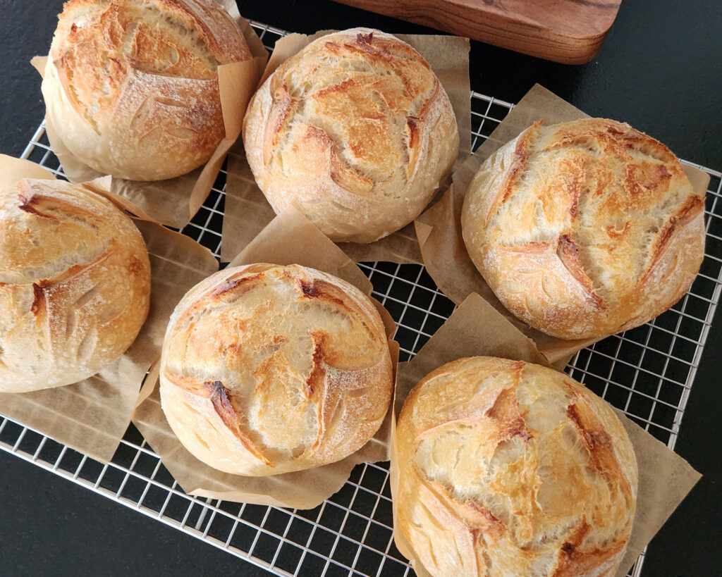 Freshly baked sourdough bread bowls cooling on a wire rack, each with a golden crust and rustic scoring, resting on squares of parchment paper.