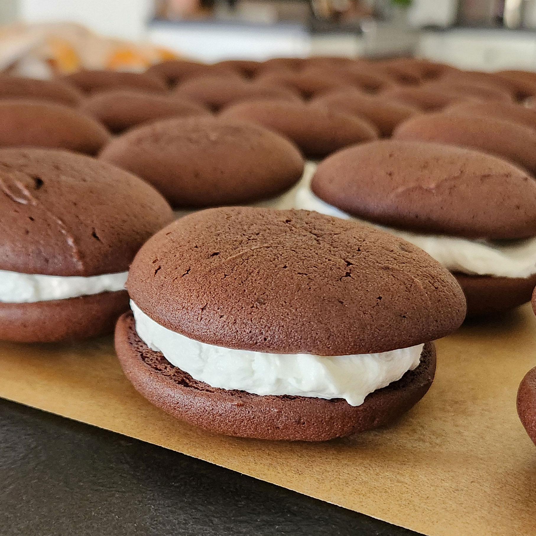Rows of freshly baked chocolate whoopie pies filled with fluffy white vanilla cream, cooling on parchment paper in a cozy kitchen setting.